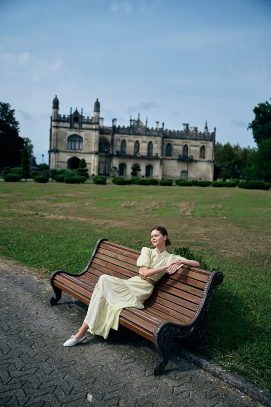 A woman sits on a wooden bench in a green park with a grand castle in the background, creating a serene outdoors fashion moment and calm scenery.
