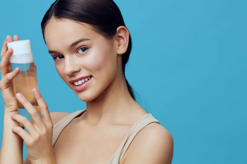 Young woman smiling with a skincare product against a blue background, promoting healthy skin and beauty concepts
