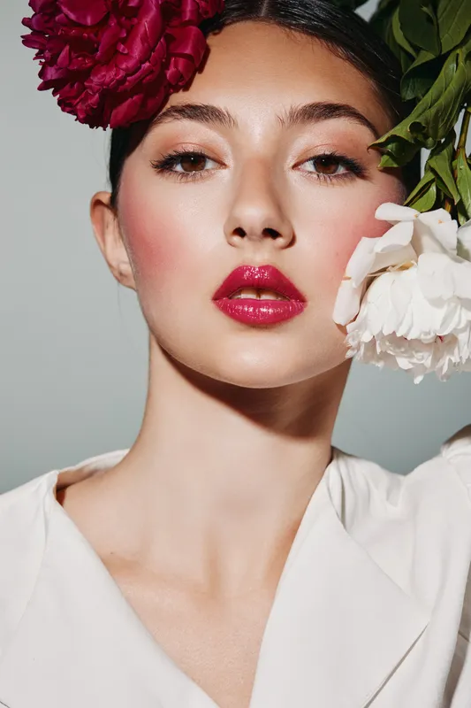 Fashion model with glossy red lips and floral headpiece poses confidently. Clean bright background highlights creative beauty and feminine elegance concept.