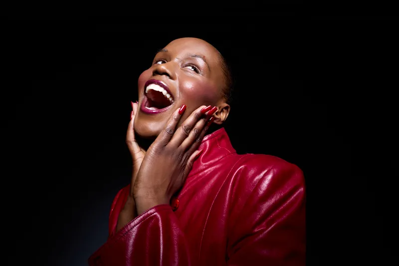 Happy African woman with dark skin wearing a red leather jacket smiles joyfully with hands on face. Black background highlights her expression of excitement and confidence in studio light.