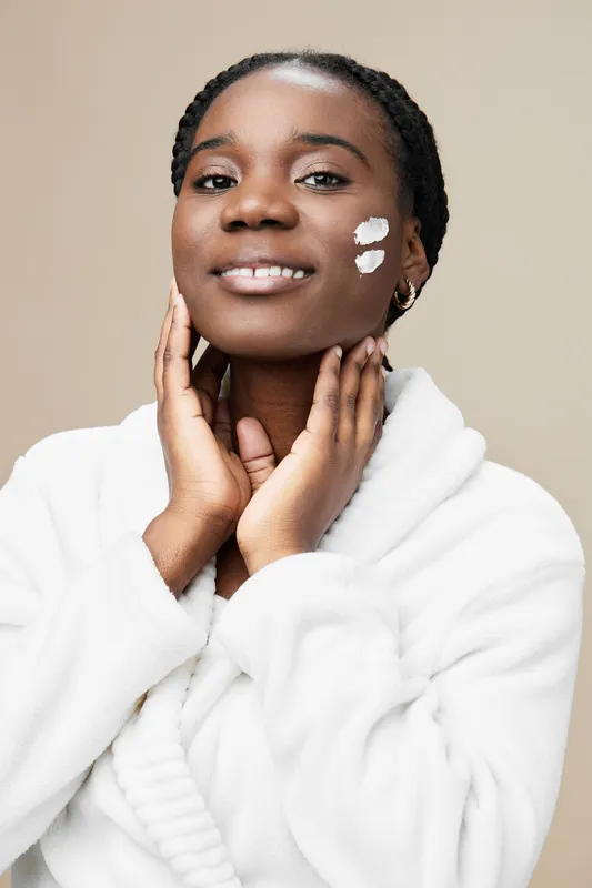 Smiling African woman applying skincare cream, wearing a cozy white robe, with a neutral beige background, showcasing a fresh and healthy skincare routine