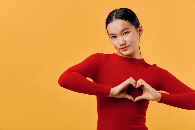 Young woman in red sweater making hand heart gesture with fingers smiling friendly on orange background. Portrait, Asian female, casual style, love symbol and positive emotion concept.
