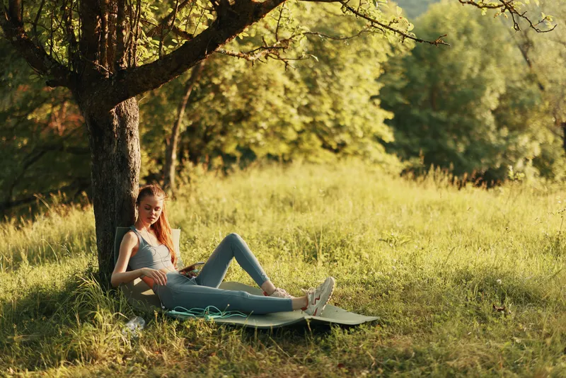 Young woman enjoying a book while resting under a tree in a green park, reflecting peace and relaxation in a natural setting