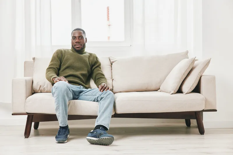 Young Black man in a cozy green sweater sitting on a modern beige sofa, relaxed and smiling, bright airy background with natural light