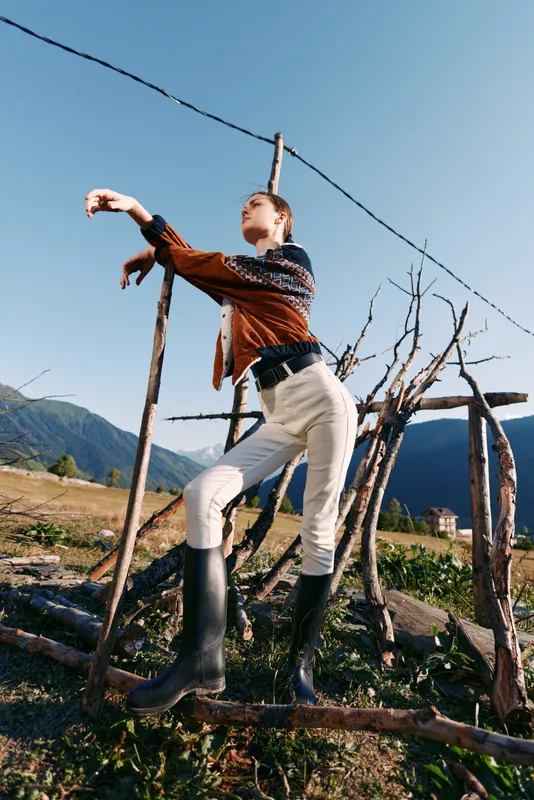 Woman model posing by rustic fence in countryside wearing rubber boots and patterned sweater, stylish outdoor fashion portrait with mountains background and confident stance