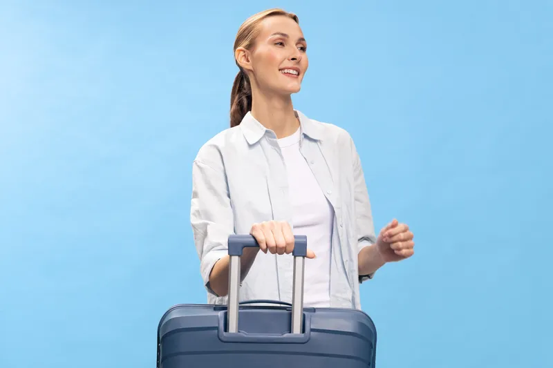 Smiling woman with suitcase looks away on blue background. Casual traveler in white shirt prepares for trip. Concept of travel, vacation, tourism, and lifestyle.