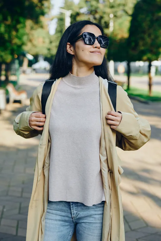 Woman in a beige coat and sweater walks outdoors in a park, confident style with sunglasses, backpack and casual pose, capturing everyday street fashion and natural light.