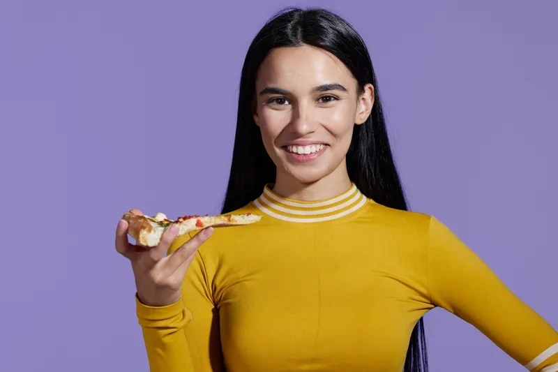 woman sport fitness with long black hair wearing yellow top smiling while holding pizza slice against purple background, healthy lifestyle concept