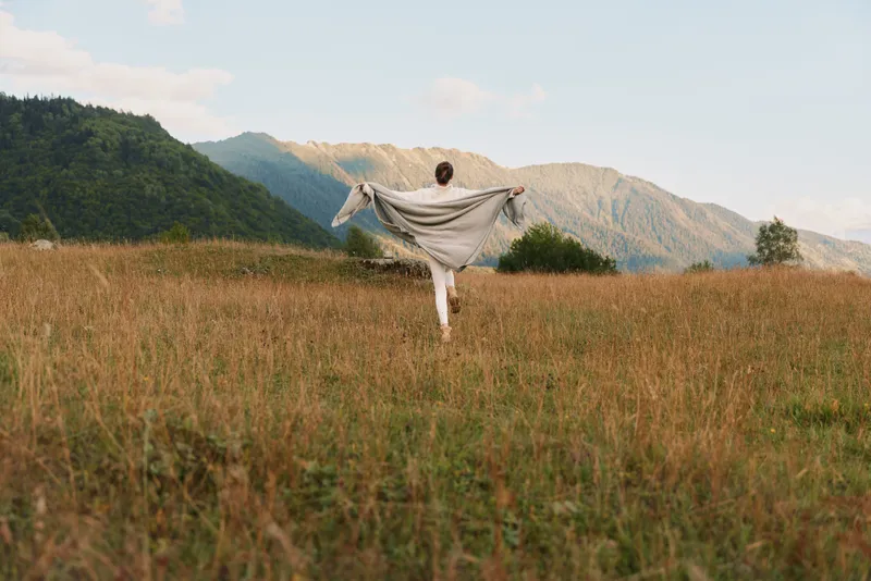 Woman in a wide meadow holds a scarf, enjoying outdoor life, embracing sunrise light among mountains, expressing freedom, calm mood, and relaxed lifestyle. Warm breeze.