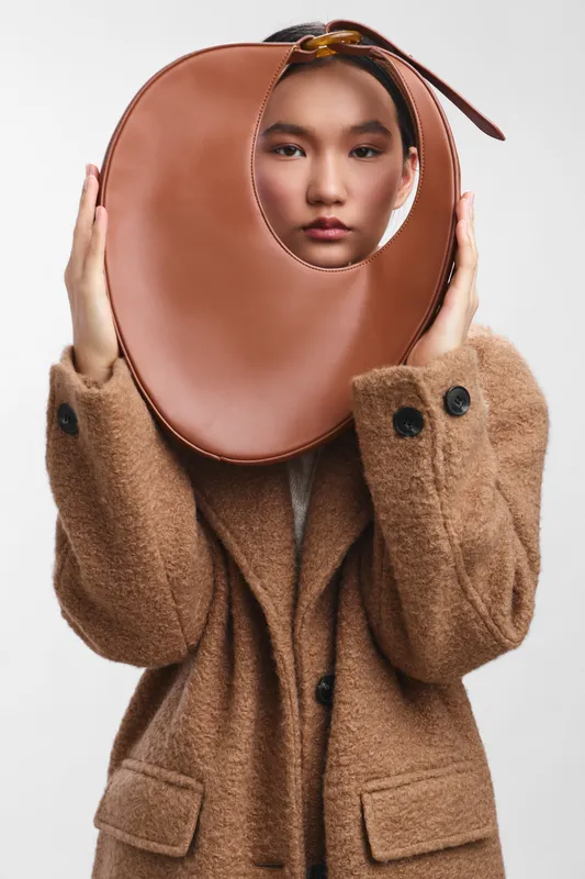 asian woman wearing a warm brown coat holds a large circular prop to her face in a studio setting, creating a playful yet stylish portrait with a contemporary fashion vibe.