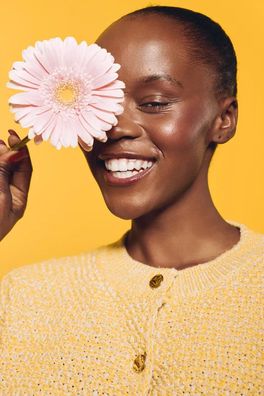 Smiling black woman with dark skin covers one eye with a pink flower. She wears a textured yellow sweater and poses against a bright yellow background. Concept of beauty and happiness.