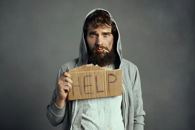 Бездомный мужчина с бородой держит табличку с надписью «Help» на улице