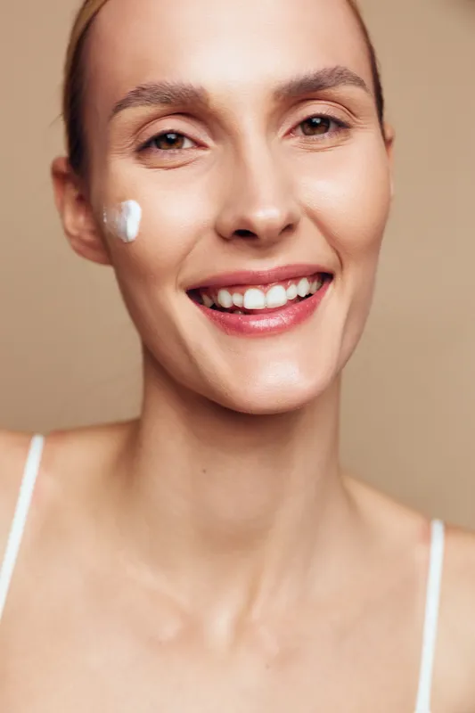 Young woman smiles while applying moisturizer on her cheek in a close up portrait, highlighting a simple skincare routine and healthy glowing skin.