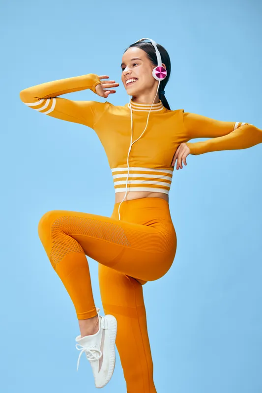 Woman in bright orange athletic outfit dances mid-air against a blue studio backdrop, wearing headphones, showcasing energetic movement, fitness lifestyle and joyful motion.