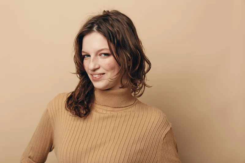 Smiling woman with wavy brown hair wearing beige turtleneck sweater looks confidently at camera on light beige background. Concept of casual style, natural beauty, and positive mood.