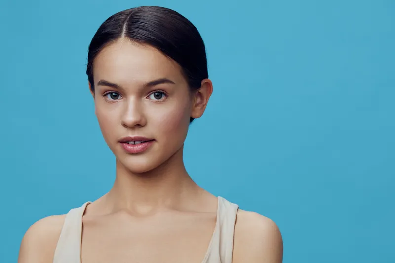 Young woman with a natural look, light clothing, and a serene expression on a blue background, embodying simplicity and beauty