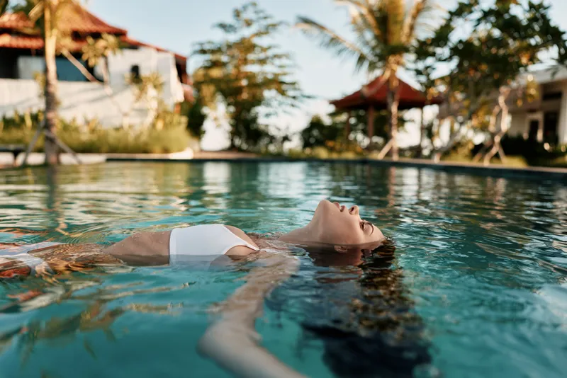 Relaxed person floating on back in a bright pool, tropical resort vibe, summer leisure and wellness under sun, calm water reflections and carefree holiday mood.
