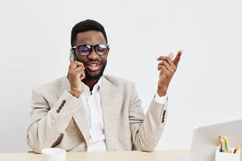 Confident young African man in beige blazer wearing sunglasses talking on mobile phone, smiling, gesturing with hand, sitting at modern desk with laptop, isolated on plain white background, studio por