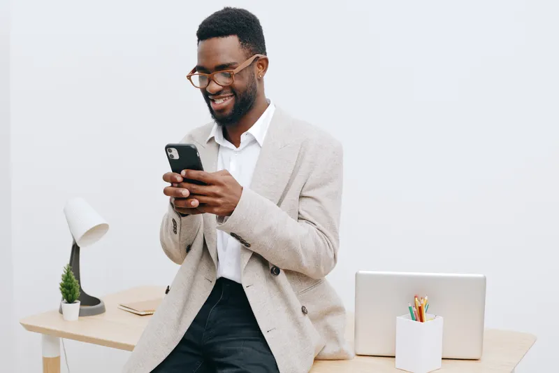 Joyful young African American man in casual office attire using smartphone looking happy smiling isolated on plain white background modern lifestyle concept