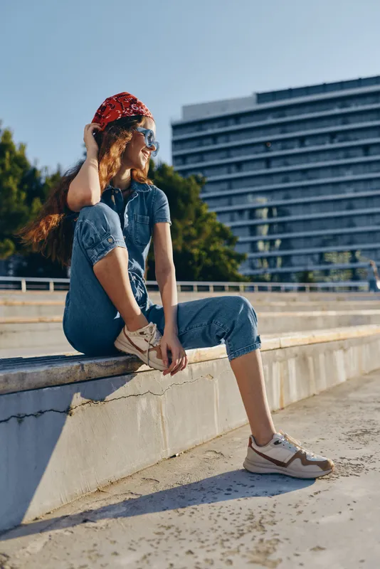 young woman sitting outdoors in casual denim outfit with red bandana and sunglasses enjoying sunny day by urban building background