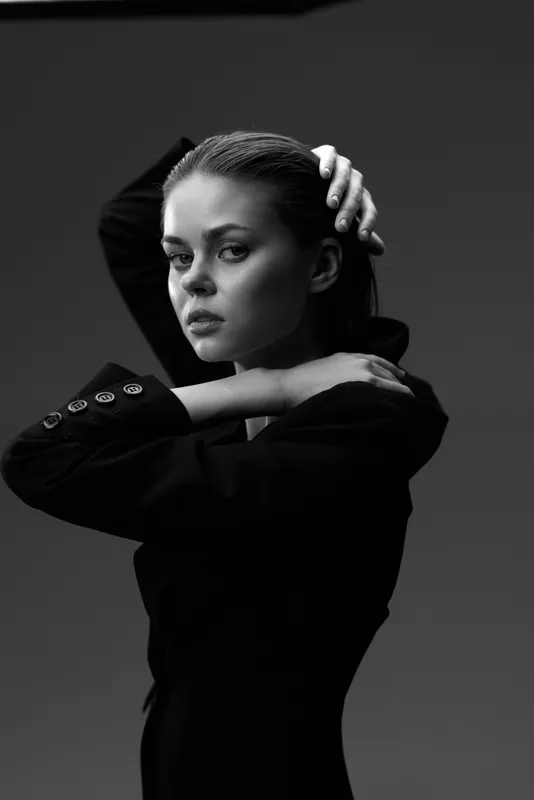 Black and white portrait of a serious woman posing with hands near head in studio lighting.