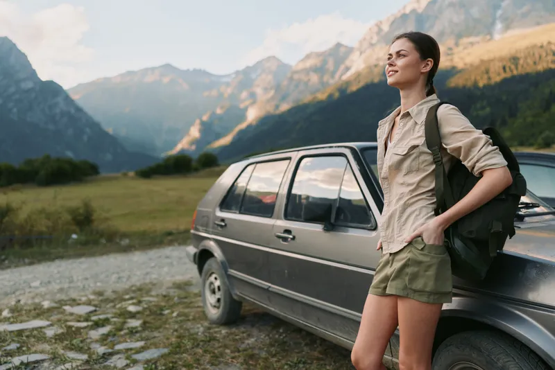 Woman with backpack by a car in a mountain landscape ready for a hike, casual outdoor attire, sunlit valley, sense of adventure and independence.