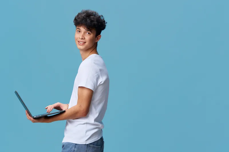 young man with laptop wearing casual white t-shirt and jeans smiling against blue background, tech, youth, cheerful, modern lifestyle