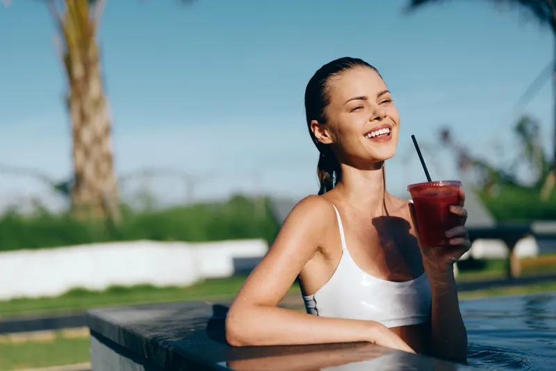 A smiling woman relaxes in a bright pool, holding a red drink. White swimsuit, sunlit water, and casual summer lifestyle convey leisure, refreshment, and radiant happiness.