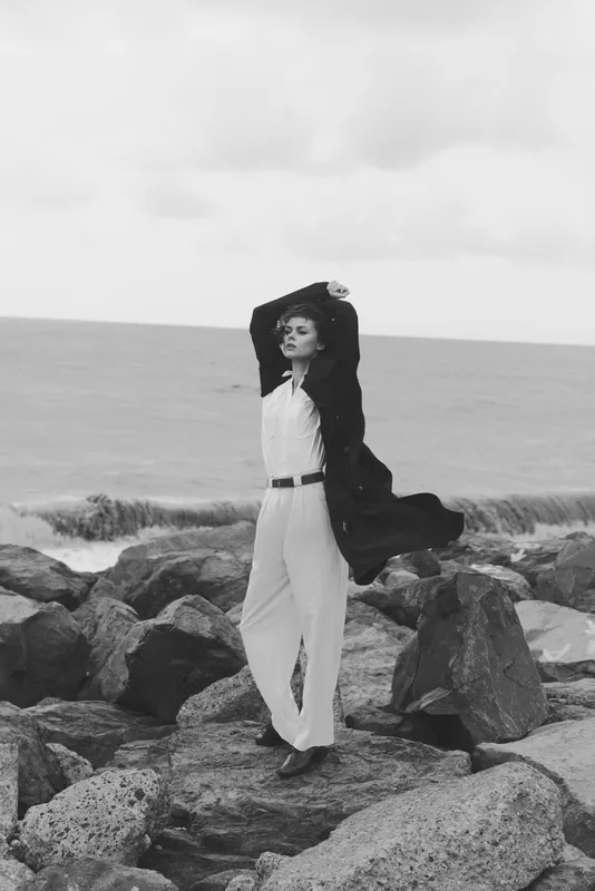 Monochrome scene of a windswept woman standing on a rocky shore in a long white dress and dark cardigan, embracing the sea breeze as waves crash behind her