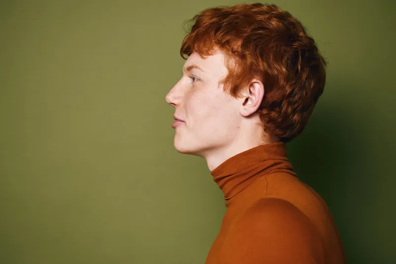 Profile of young man with red hair and freckles wearing brown turtleneck standing against green background in studio. Side view portrait with calm expression and casual outfit for fashion.