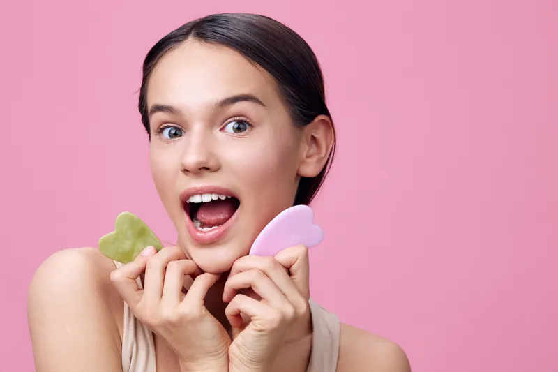 Joyful model holding facial massage tools against a pink background, promoting skincare and wellness concepts, expressing excitement and freshness