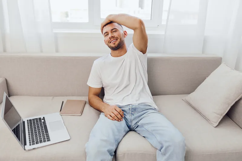 Relaxed man enjoying leisure time at home, sitting on a sofa with a laptop and notebook. Bright and airy atmosphere enhancing comfort and tranquility.