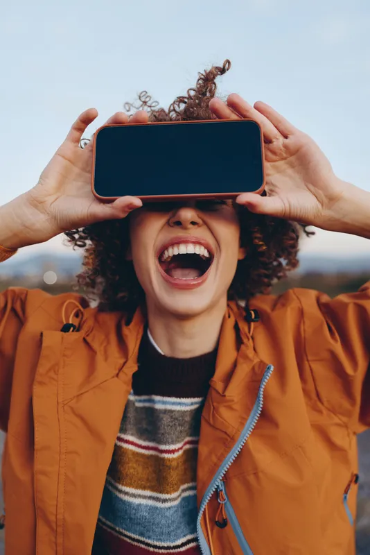 Woman lifestyle fun outdoors, laughing with hands holding a smartphone, curly hair on her face, street background bokeh, joyful candid moment for mobile media, sharing video now