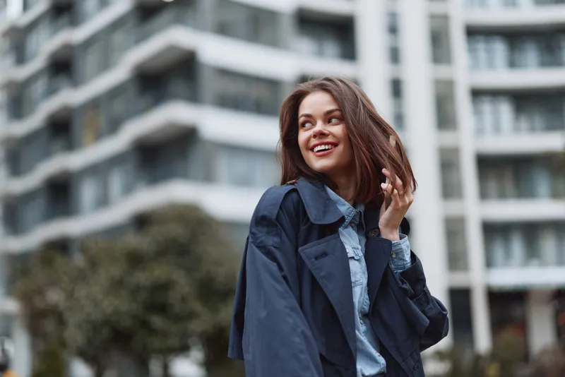 Young woman with long brown hair smiles happily while standing outdoors in a casual blue jacket and denim shirt in an urban environment with blurred buildings behind her.