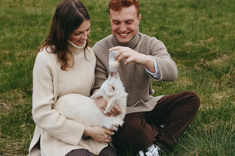 Young couple sitting on grass feeding a white baby goat with a bottle enjoying a cozy outdoor lifestyle moment in warm sweaters during autumn season.