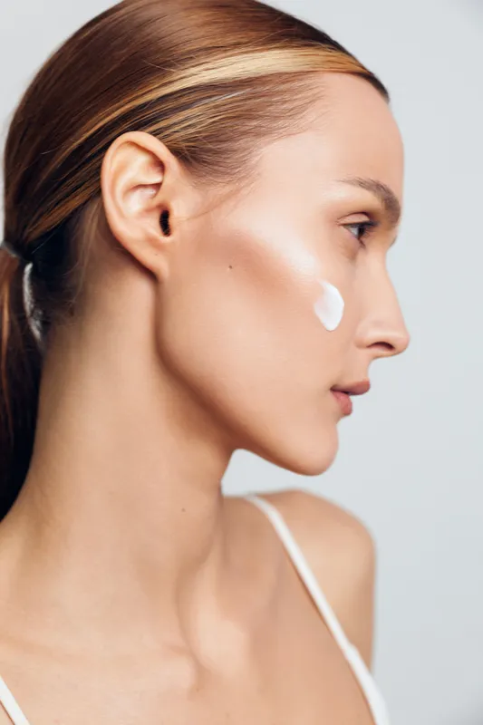 Young woman with clear skin applies moisturizing cream on her cheek in profile view isolated on white background with natural makeup and sleek hair tied back in ponytail