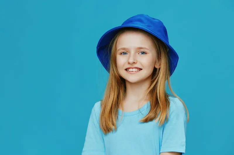 Cheerful young girl with blonde hair wearing a blue hat and a light blue shirt on a vibrant turquoise background, radiating joy and happiness