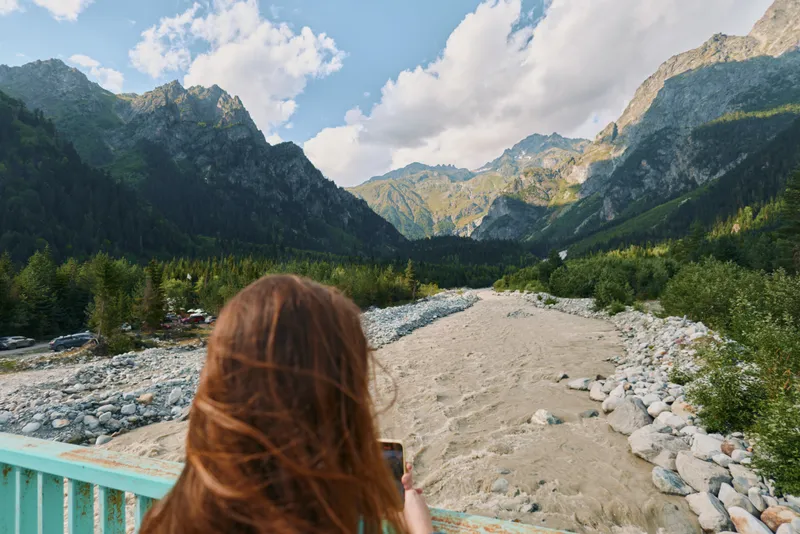 A traveler stands on a bridge and photographs a wide river valley framed by towering mountains. The scene conveys adventure, nature, and quiet exploration.