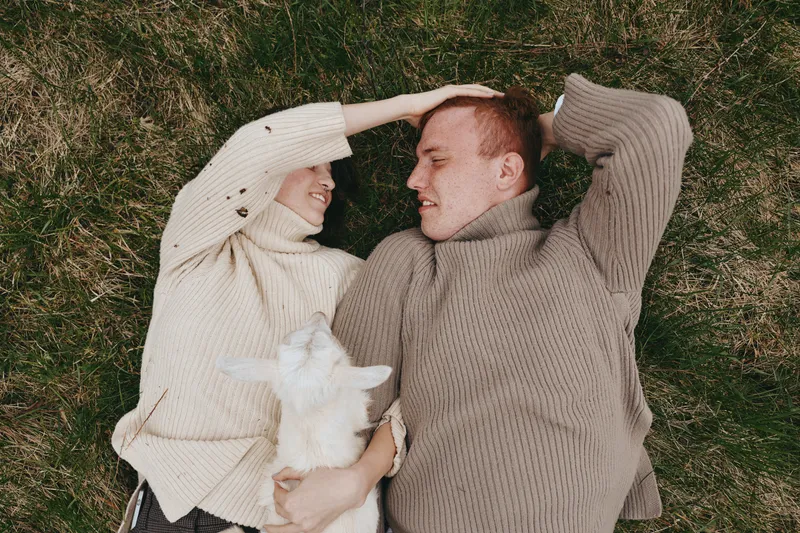 Loving couple in cozy sweaters lie on grass with a playful goat kid between them, capturing rustic lifestyle, affection, and animal companionship in soft daylight.