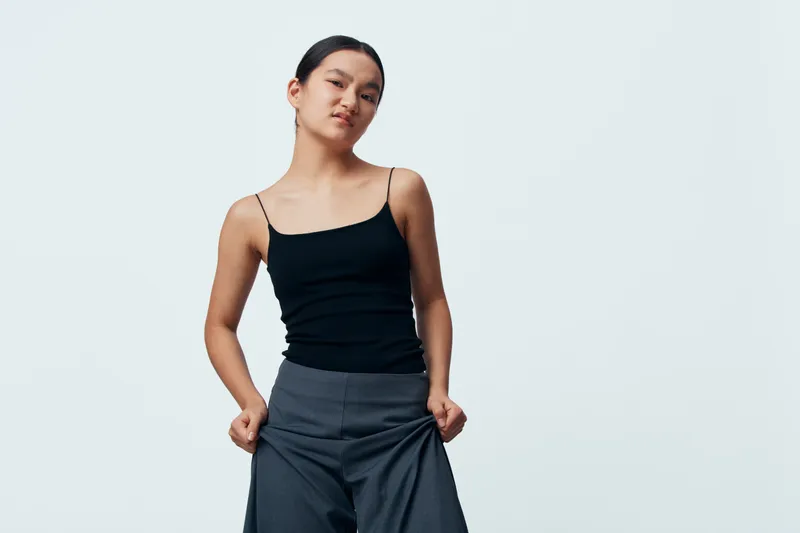 Young woman in black tank top and gray pants posing confidently with hands in pockets on light background. Casual fashion style, confident female model standing with relaxed posture.