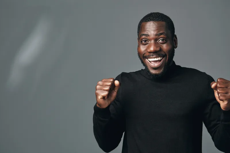 African man with dark skin wearing black sweater smiling and celebrating success with clenched fists in creative conceptual visual style on gray background.