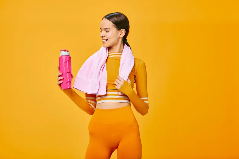 Fitness driven woman in a vibrant athletic outfit with a towel around her neck, holding a pink water bottle against a bold orange backdrop, ready for workout and hydration.
