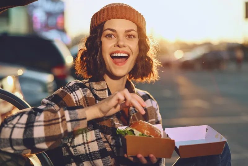 Woman portrait with a bright smile holding a burger takeout outdoors in an urban sunset, candid moment capturing authenticity, mindful living and golden hour glow with emotional storytelling.