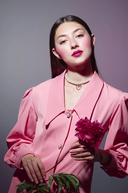 Young woman in bright pink vintage style blouse with minimal makeup and dark hair holds a peony flower. Fashion portrait with creative beauty concept and soft studio lighting.
