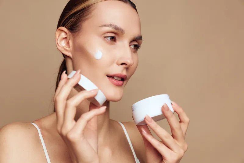 Young woman with blonde hair applies moisturizer on her cheek during a skincare routine, in a beige studio setting, with calm, focused expression and dewy skin.