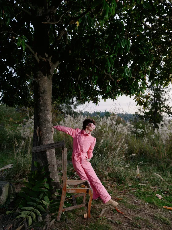 Person in a pink jumpsuit leaning on a wooden chair under a leafy tree in a sunny outdoor setting, showcasing relaxed casual fashion and natural landscape.