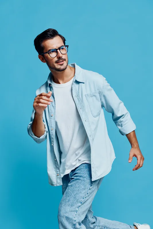 Stylish happy man wearing glasses and light blue denim shirt walking confidently while holding a pen, set against a vivid blue background. Lifestyle concept.