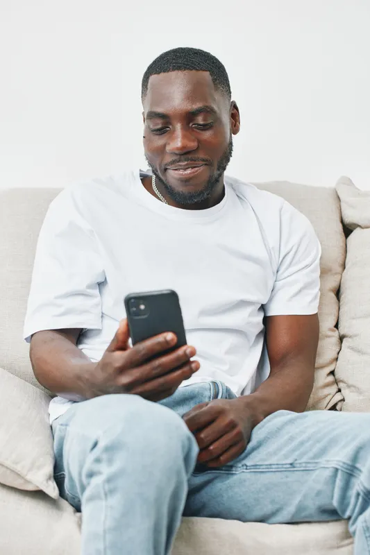 Young Black man smiling while using smartphone on couch, wearing casual outfit, capturing moments of joy and relaxation at home