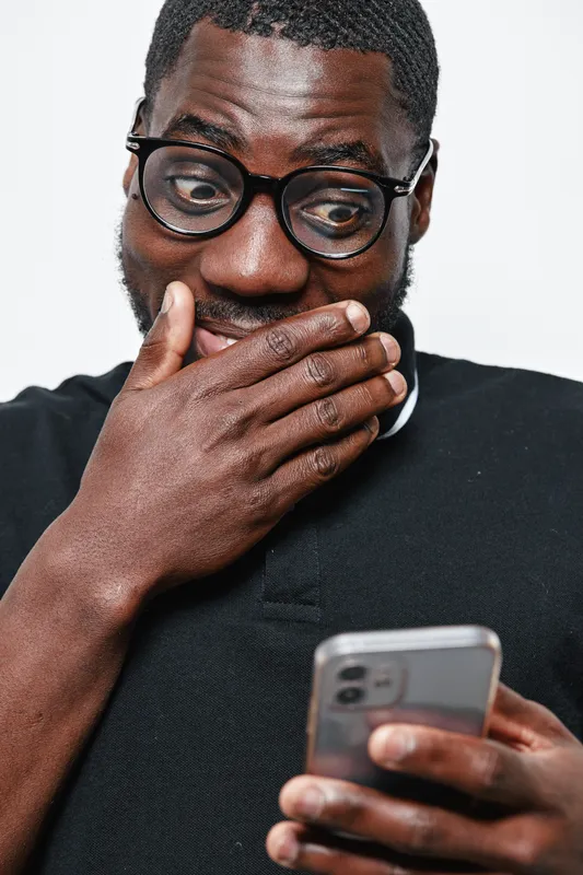 Black man with dark skin and glasses looking surprised while holding smartphone and covering mouth with hand. Casual clothing, technology, emotion and communication concept.