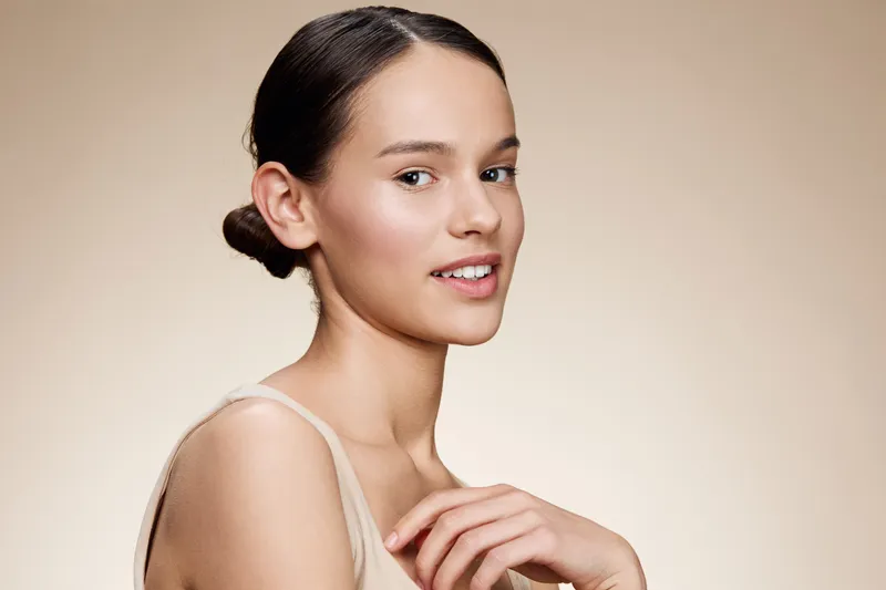 Young woman with natural makeup and a gentle smile, posing against a soft beige background showcasing beauty and confidence in a modern, minimalistic style